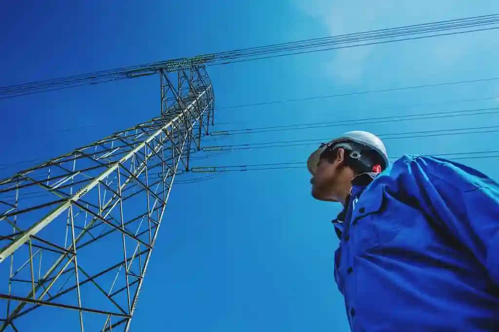Toza moviy osmon qarshisida elektr ustuni yonida turgan past burchakli muhandis | Low angle view engineer standing by electricity pylon against clear blue sky