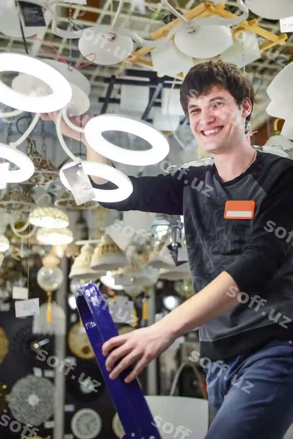 Do'kon xodimi chiroq fonida zinapoyada jilmayib turibdi | Employee chandelier store stands stepladder smiles against background lamps