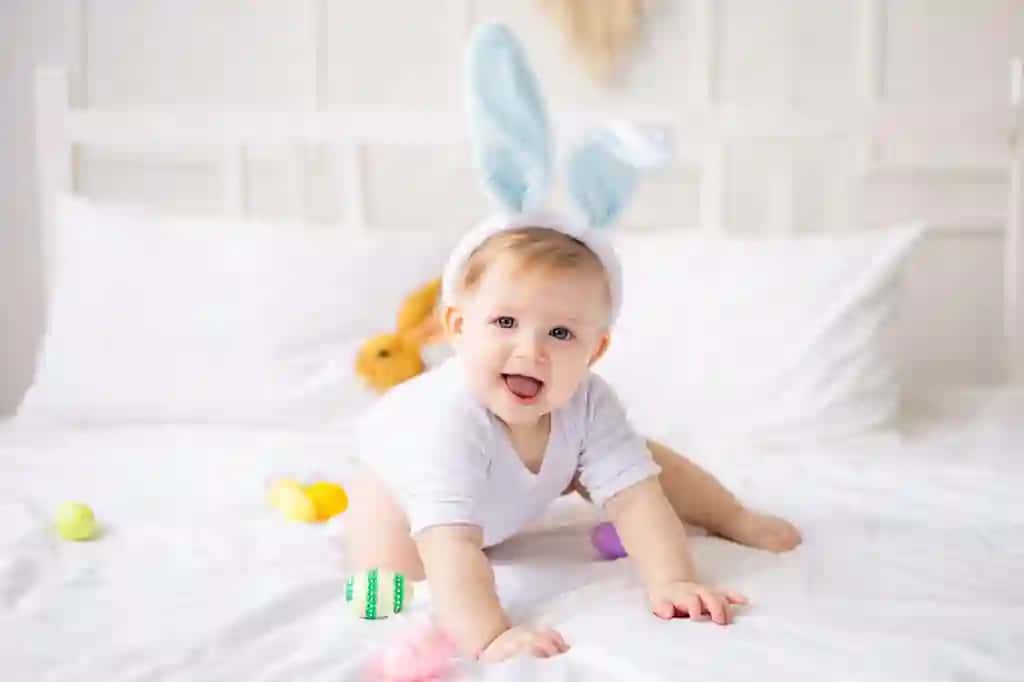 Yoqimli esnayotgan bolakay quyon quloqlari bilan rang-barang tuxum oppoq karavot uyida sarg‘ish sochli go‘dak bilan o‘ynamoqda. | Cute yawning baby boy with bunny ears colorful eggs white bed home playing little blonde baby with rabbit happy easter concept
